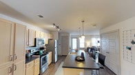 This single-floor residence offers a thoughtfully designed open-concept layout that seamlessly connects the kitchen, dining, and living areas. The kitchen is well-equipped with stainless steel appliances, including a refrigerator, electric range, and over-the-range microwave, all complemented by light wood cabinetry and dark countertops. A central island with a built-in sink and dark countertop provides additional workspace and doubles as a casual dining area with bar-height seating. Overhead, a combination of track lighting and elegant pendant lights illuminate the space, adding both functionality and style. The flooring throughout features warm-toned hardwood, lending a cohesive and inviting feel to the open floor plan. Natural light filters in through windows at the far end of the space, brightening the living area and highlighting the neutral color palette of white and grey tones on the walls. The living area, visible from the kitchen, offers a comfortable and airy atmosphere. Overall, the space balances practicality with modern aesthetics, making it an ideal environment for both everyday living and entertaining.