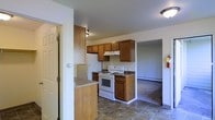 This single-floor space centers around a practical kitchen equipped with warm honey-toned wood cabinetry spanning both upper and lower configurations, providing ample storage. The kitchen features white appliances, including a freestanding electric range with overhead ventilation and a full-sized refrigerator. Granite-style countertops add a touch of visual interest to the workspace. A peninsula counter extends into the space, offering additional prep area and a natural divider between the kitchen and the rest of the unit. The flooring in the kitchen consists of durable tile in a neutral stone pattern, lending a grounded aesthetic to the area. A doorway from the kitchen leads to an adjacent room, likely a bathroom or utility space, while an open passage connects directly to a carpeted living area. The living space features baseboard heating and neutral wall tones throughout, creating a consistent and cohesive look. Flush-mount ceiling lighting provides overhead illumination, and the overall layout is straightforward and efficient, making good use of the available floor space on this single level.