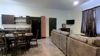 This single-floor space is designed with an open-concept layout that efficiently combines the living, dining, and kitchen areas into one cohesive environment. The living area is furnished with a large, comfortable sectional sofa in a warm brown tone and a wall-mounted flat-screen television, creating a practical and inviting setting for relaxation. The polished concrete flooring runs throughout the space, lending it an industrial yet clean aesthetic that is easy to maintain. The kitchen is equipped with dark cabinetry, a full-size black refrigerator, and open shelving, offering functional storage and workspace. Adjacent to the kitchen, a dining area features a wooden table paired with matching X-back chairs, providing a casual spot for meals. The space is illuminated by a ceiling fan with a built-in light fixture, contributing to a well-lit and comfortable atmosphere. Light-colored walls keep the interior feeling open and airy, while a dark front entry door provides a bold contrast. Overall, this space offers a straightforward and functional layout suited to everyday living, with durable finishes and practical furnishings throughout.