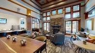 Located in Colorado Springs, CO, this single-story community clubhouse on Snow Creek Point is a masterfully designed gathering space that blends mountain-lodge aesthetics with modern amenities. The grand main lounge commands attention with soaring double-height ceilings, rich wood-trimmed windows arranged in dramatic stacked rows, and a floor-to-ceiling stacked-stone fireplace as the central focal point. Comfortable seating arrangements invite relaxed conversation, while an adjacent nook with a sofa and café table offers a more intimate retreat bathed in natural light from French doors opening to the outdoors.

The welcoming entry foyer features light hardwood-style flooring, warm stone-clad columns, and wood millwork details that establish the craftsmanship found throughout. A well-appointed kitchenette provides sleek dark cabinetry, white countertops, stainless steel appliances, and a wall-mounted display screen. A game and activity area includes a shuffleboard table and chess table set against large windows framing views of the surrounding Colorado landscape. A dedicated lounge corner with mounted televisions, wood wainscoting, and lush greenery adds a casual, social atmosphere. Together, these spaces form a cohesive, inviting clubhouse designed for community connection.