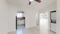 This single-floor studio in Miami Beach's South of Fifth neighborhood offers a bright, open layout finished in crisp white throughout. Large-format light gray tile flooring runs seamlessly across the main living area, complemented by high ceilings and ceiling fans that enhance airflow and comfort in the Florida climate. A charming arched doorway frames the kitchen, which features white shaker cabinetry, warm tile backsplash, stainless steel appliances including a refrigerator and gas range, a built-in sink, and a wall-mounted A/C unit with a window for natural light. The spacious main living area benefits from generous corner windows that flood the room with sunlight and offer lush, tree-lined outdoor views. The bathroom is neatly appointed with neutral stone-tiled floors, full-wall tile in the glass-enclosed walk-in shower, and a window for ventilation. A dedicated area includes an in-unit stacked washer and dryer alongside a built-in closet with shelving, providing practical storage. The entry area features polished concrete-style flooring and a smart home panel. The overall ambiance is clean, minimalist, and airy—well-suited for the vibrant Miami Beach lifestyle.