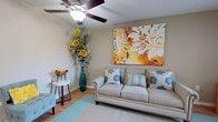 This single-story residence offers a thoughtfully designed layout with distinct living spaces connected by warm hardwood-style flooring throughout the main areas. The living room serves as a central gathering space, featuring a ceiling fan, neutral walls, and a large, vibrant abstract canvas above a classic rolled-arm sofa. Decorative accents, including tall floral arrangements and patterned throw pillows, add personality to the room.

Adjacent to the living area, the dining space accommodates a dark wood table with upholstered seating beneath a pendant light fixture, complemented by colorful wall art and a lush potted plant. A separate flex room offers additional versatility, bathed in natural light through wide windows dressed with sheer dark curtains.

The kitchen is equipped with stainless steel appliances, including a refrigerator and over-range microwave, alongside light wood cabinetry and tile flooring. Two full bathrooms feature tub-shower combinations, light wood vanities with granite-style countertops, and tile flooring. The bedroom includes a ceiling fan and a walk-in closet with built-in shelving. A dedicated laundry area with a full-size washer and dryer and overhead shelving completes the unit.