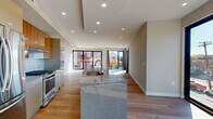 This contemporary single-floor residence showcases a thoughtfully designed open-concept layout that seamlessly connects the kitchen, dining, and living areas. The kitchen is a standout feature, centered around a large granite-topped island with an integrated sink and high-arc faucet. Cabinetry combines rich warm-toned wood uppers with sleek grey lower units, complemented by a subway tile backsplash and stainless steel appliances including a gas range and refrigerator. Recessed lighting and a distinctive coffered ceiling detail above the kitchen add architectural depth.

Wide-plank hardwood flooring flows throughout the space, unifying each area with warmth and continuity. The living area is bright and expansive, benefiting from floor-to-ceiling black-framed windows and sliding glass doors that open to a private balcony, inviting generous natural light and outdoor views of the surrounding neighborhood.

The bedrooms are well-proportioned with neutral tones and continued hardwood flooring. The bathrooms reflect a refined modern aesthetic — one features a wall-mounted trough sink with a dark wood vanity, while another offers a frameless glass walk-in shower with textured tile surround and mosaic pebble flooring. Every detail of this residence reflects clean, contemporary design with quality finishes throughout.