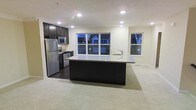 This 3-floor space showcases a thoughtfully designed open-concept layout, with the kitchen and living area seamlessly flowing together on the main level. The kitchen is outfitted with dark espresso cabinetry that creates a striking contrast against the light-toned countertops. A generously sized central island anchors the space, featuring an integrated sink and providing ample workspace and casual dining potential. Stainless steel appliances, including a refrigerator, microwave, and range, lend a sleek, contemporary feel to the kitchen. Recessed lighting throughout the ceiling ensures consistent, well-distributed illumination across the space. Multiple windows along the exterior wall allow natural light to filter in during daytime hours while offering views of the surrounding neighborhood. The transition from dark hardwood flooring in the kitchen to neutral-toned carpet in the adjacent living area subtly defines each zone while maintaining an open, cohesive feel. The light-colored walls and clean architectural lines contribute to a bright, modern ambiance throughout. With three floors in total, this space offers substantial room for varied living arrangements across its multiple levels.