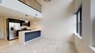 This two-story loft showcases a striking contemporary design defined by soaring double-height ceilings in the main living area. The ground floor is anchored by a spacious open-plan living and kitchen zone finished with polished concrete flooring that reflects light throughout the space. Large black-framed windows line the living area, framing urban views and flooding the interior with natural light.

The kitchen is thoughtfully appointed with dark cabinetry, stainless steel appliances including a gas range, microwave, and French door refrigerator, and a two-tone peninsula featuring a crisp white countertop and navy blue base. Pendant lighting hangs above the peninsula, adding a modern, industrial touch.

A floating staircase with a black metal railing and warm wood treads connects the two levels, leading to an upper mezzanine that overlooks the main living space below. The second floor features warm hardwood flooring, providing a contrast to the polished concrete downstairs.

The full bathroom presents white hexagonal tile surround walls, a soaking tub, dark slate-toned floor tile, and a floating dark vanity. A separate powder room on the main level offers a clean, minimalist design. Overall, the space delivers an urban loft aesthetic with refined, modern finishes throughout.