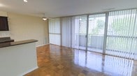 This single-floor space is defined by its open layout and abundance of natural light. The main living area showcases polished parquet flooring laid in a classic block pattern, lending warmth and character to the room. A striking wall of floor-to-ceiling sliding glass doors, dressed with vertical blinds, spans the exterior wall, flooding the interior with daylight and providing direct views of lush greenery and the surrounding outdoor environment. The glass doors open to what appears to be a balcony or exterior walkway, seamlessly connecting the indoors with the outside. A ceiling fan is centrally positioned, offering both practical ventilation and a casual aesthetic touch. One wall features a textured surface, adding visual contrast to the otherwise smooth finishes throughout the space. A partial countertop or partition is visible near the edge of the frame, suggesting a defined transition between the living area and an adjacent kitchen or utility space. The overall ambiance is clean, luminous, and functional, with the polished floors and expansive glazing working together to create a sense of openness and continuity throughout the interior.