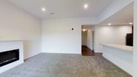 This single-floor space offers a thoughtfully designed layout that balances openness with functionality. The main living area is finished with plush gray carpeting that adds warmth and comfort underfoot, while crisp white walls create a bright, neutral backdrop that can accommodate a variety of interior styles. Recessed lighting in the ceiling provides clean, even illumination throughout the space.

A wide opening connects the living area to a hallway, which leads to additional rooms beyond, hinting at a well-organized floor plan. The transition from carpet to dark hardwood-style flooring in the hallway adds a visual distinction between the living and private areas of the unit. A partial countertop or shelf is visible along one wall, suggesting a convenient pass-through or breakfast bar feature that adds practical utility to the layout.

The overall ambiance is clean, modern, and move-in ready, with a minimalist aesthetic defined by smooth ceilings, neutral tones, and consistent finishes throughout. The space's simplicity makes it highly versatile, offering a blank canvas that can be personalized to suit a range of lifestyles and furnishing preferences.
