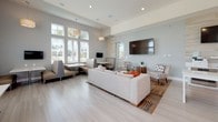 This single-floor communal space is designed with a contemporary, coastal-inspired aesthetic that balances functionality with comfort. The open-concept layout seamlessly connects several distinct zones, creating an inviting and versatile environment throughout.

The kitchen area is a standout feature, anchored by a long, white quartz island with an integrated microwave and generous counter space. White shaker cabinetry, pendant lighting, and a stainless steel refrigerator contribute to a clean, polished look. A dramatic glass-paneled garage-style door opens the kitchen to the outdoors, flooding the space with natural light and providing a strong visual connection to the exterior. An adjacent countertop bar with white corbel supports and woven rattan barstools offers a casual seating option, complemented by dual wall-mounted televisions and a tiled accent wall.

The lounge area features a plush neutral sectional sofa, accent chairs with geometric throw pillows, and a sculptural round coffee table, all arranged atop a richly patterned area rug. A large wall-mounted television serves as a focal point. Booth-style dining nooks with upholstered banquette seating and white pedestal tables line the perimeter near tall windows, which frame views of lush greenery and palm trees. Light wood-look flooring and soft gray walls unify the entire space with an airy, relaxed ambiance.