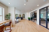 Located on Presidential Way in West Palm Beach, Florida, this single-floor leasing office offers a thoughtfully designed space that blends modern aesthetics with warm, welcoming details. Light hardwood flooring runs throughout, complementing the crisp white walls and recessed lighting that keep the interior bright and airy. At the heart of the space is an elegant reception desk finished in brushed steel with a white countertop, illuminated by a trio of globe pendant lights with gold accents. Behind the desk, arched alcoves adorned with lush botanical wallpaper in rich greens and golds create a striking focal point that infuses the space with a tropical Florida character. Private offices are enclosed by sleek black-framed glass partition walls, offering visual openness while maintaining a sense of privacy for staff. The lounge area features plush rounded seating arranged around an ornate area rug, bathed in natural light from large windows that frame views of the surrounding greenery. A standing work counter with bar-height seating provides a flexible workspace. A separate area with mailboxes and additional seating completes the functional layout. Potted tropical plants are strategically placed throughout, reinforcing the fresh, nature-inspired atmosphere of this well-appointed leasing office.