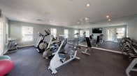 Located at 734 S Mesa Hills Dr in El Paso, Texas, this dedicated fitness center offers a well-equipped, single-floor space designed to support a comprehensive workout routine. The room features cool-toned, light gray walls paired with dark, multi-toned carpet tiles that lend the space a clean, modern aesthetic. Recessed lighting and natural light from multiple windows create a bright and energizing atmosphere, with views of surrounding greenery adding a pleasant outdoor connection.

The facility is outfitted with a diverse selection of cardio equipment, including a treadmill, elliptical trainer, and stationary recumbent bike, all positioned to allow comfortable use throughout the space. A dedicated free weights area features a full dumbbell rack alongside an adjustable weight bench, catering to strength training needs. A large wall-mounted mirror spans one side of the room, enhancing the sense of space and allowing users to monitor their form during workouts. A wall-mounted flat-screen TV provides entertainment options during exercise sessions.

Additional amenities include a water fountain for convenient hydration, a designated door entry with glass paneling that connects to the exterior, and proper safety provisions including a fire extinguisher. The open floor plan allows for versatile use of the space, accommodating both individual and group fitness activities with ease.