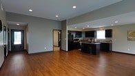 This single-floor space showcases a thoughtfully designed open-concept layout that seamlessly connects the living and kitchen areas. The warm-toned hardwood flooring runs throughout, anchoring the space with a sense of continuity and natural warmth.

The kitchen is a standout feature, outfitted with sleek dark cabinetry that extends to the ceiling, providing ample storage. A central island with bar-height seating serves as both a functional workspace and a casual dining spot. Full-sized appliances, including a refrigerator and built-in microwave, are integrated into the cabinetry for a streamlined look. A window above the sink allows natural light to brighten the workspace.

Recessed lighting is installed throughout the ceiling, offering even, adjustable illumination across the open floor plan. The walls are finished in a neutral gray tone, complementing the contrast between the dark kitchen elements and the light ceiling.

A hallway visible from the main living area leads to additional rooms, suggesting a practical and well-organized floor plan. The generous open living area offers flexibility for a variety of furniture arrangements, making the space both versatile and inviting. Overall, the design balances modern aesthetics with everyday functionality.