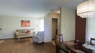 This two-floor residence offers a thoughtfully designed layout that prioritizes comfort and functionality. The main living area features rich, dark hardwood-style flooring that extends throughout, creating a cohesive and warm aesthetic. The open-concept arrangement allows the dining and living spaces to flow naturally into one another, making the area feel spacious and inviting.

The living room is well-lit, drawing in natural light through a window that brightens the neutral-toned walls. The space comfortably accommodates a seating arrangement, including a sofa and accent chair, complemented by a glass-top coffee table. A vibrant piece of wall art adds a pop of color against the light, neutral palette.

Adjacent to the living area, a doorway reveals a closet with a built-in rod, offering convenient storage. A wall-mounted thermostat provides easy climate control. The dining area, positioned nearby, features a dark wood table and chairs, anchored by a patterned area rug. Together, the spaces present a practical and welcoming interior that carries throughout both floors of the residence.