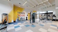 This contemporary fitness center is designed to inspire and energize. The open, single-floor layout is anchored by a bold motivational mural — "Train Insane or Remain the Same" — splashed across a vibrant yellow accent wall, immediately setting an energetic tone for every workout session. The space features a distinctive checkered floor pattern in blue, beige, and gray tones that adds visual dynamism while remaining practical and durable underfoot. Overhead, sleek linear pendant lighting complements recessed fixtures to ensure the space is consistently well-lit throughout the day. Large floor-to-ceiling windows along multiple walls flood the interior with abundant natural light and offer views of the surrounding outdoor amenity areas, including what appears to be a pool deck. The cardio zone is equipped with treadmills, elliptical trainers, and a rowing machine, all positioned to take advantage of the outdoor views. A cable machine and multi-use strength station serve as the centerpiece for resistance training, while a dedicated free weights section features a full dumbbell rack paired with an adjustable bench. A separate stretching and functional training area provides ample open floor space alongside a storage rack stocked with medicine balls and resistance accessories, making this fitness center a comprehensive and inviting facility.