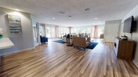 This generously proportioned single-floor space is designed with an open-concept layout that seamlessly connects multiple functional areas. The expansive main living area features warm wood-look flooring, recessed lighting, and a textured ceiling, creating a bright and welcoming atmosphere. Large windows and glass doors line the perimeter, flooding the interior with natural light and offering views of lush outdoor greenery. The living area is furnished with comfortable seating arrangements, including a sectional sofa, accent chairs, and a media console with a wall-mounted television, making it well-suited for both relaxation and gathering.

Adjacent to the living area, a dedicated dining space accommodates multiple seating arrangements with wood-and-teal chairs positioned alongside floor-to-ceiling windows that frame verdant outdoor scenery. The kitchen is well-appointed with grey cabinetry, white countertops, stainless steel appliances including a refrigerator and dishwasher, and a pass-through counter that enhances connectivity to the broader space. A circular decorative mirror accent and carefully curated artwork add visual interest throughout. The overall ambiance is modern, light-filled, and versatile, suitable for a variety of communal or multi-purpose uses.