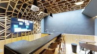 This single-floor space makes an immediate impression through its standout architectural ceiling, which features an elaborate geometric lattice framework crafted from natural wood. The interlocking angular patterns span the entire overhead space, creating a visually dynamic canopy that draws the eye upward and adds considerable depth and texture to the interior.

The space centers around a long, dark matte countertop that serves as both a functional workspace and a social gathering surface. A row of wooden bar stools lines one side, offering casual seating with a warm, natural finish that complements the wood tones of the ceiling. The countertop extends to a built-in sink area, indicating utility and convenience within the same open layout.

Lighting is a key design element, with a mix of black conical pendants and woven rattan-style pendant lights suspended at varying heights from the lattice ceiling. This combination balances industrial and organic aesthetics effectively.

A large mounted display screen is integrated into the wood lattice wall on one side, suggesting the space is designed for presentations or collaborative use. The cool blue-grey walls provide a neutral contrast to the warmth of the natural wood, resulting in a cohesive, modern, and purposefully designed interior.