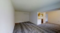 This single-floor space offers a practical and straightforward layout designed for comfortable everyday living. The main living area is spacious and open, finished with wall-to-wall carpeting in a neutral grey tone that adds a soft, understated feel underfoot. White walls throughout keep the interior bright and versatile, providing a clean backdrop ready for personalization.

A built-in closet with sliding panel doors lines one wall of the living area, offering convenient and accessible storage without occupying additional floor space. The seamless white finish of the closet doors blends naturally with the surrounding walls.

A pass-through opening connects the living area to the kitchen, promoting an open, communicative flow between the two spaces. The kitchen features warm-toned wood cabinetry along the upper walls, providing ample storage, while a countertop peninsula at the pass-through serves as both a workspace and an informal divider. The kitchen floor transitions to a hard surface, contrasting with the carpeted living area and making for easy maintenance.

Overall, the space presents a functional, move-in-ready environment with a simple, clean aesthetic suited to a range of occupants.
