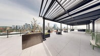 Located atop 250 Rue Young in Montréal's dynamic urban core, this expansive rooftop terrace offers a sophisticated outdoor retreat designed with contemporary elegance. The terrace is defined by a striking steel pergola structure that runs along much of its length, providing partial shade while framing sweeping panoramic views of the Montréal skyline. Large-format light grey porcelain pavers cover the entire floor, lending the space a clean and refined aesthetic.

Substantial dark metal planter boxes are distributed thoughtfully across the terrace, filled with greenery, small trees, and decorative stone, introducing natural elements that contrast beautifully with the modern industrial design. Fire features integrated into sleek metallic planters serve as focal points, adding warmth and ambiance. Seating areas are arranged throughout, including woven green lounge chairs under the covered pergola section and modular sofas positioned to take full advantage of the cityscape views.

A partially enclosed zone beneath the pergola offers a sheltered gathering area with white paneled walls and built-in track lighting overhead, suitable for events in variable weather. The open-air sections of the terrace capture unobstructed views of the Montréal skyline. The overall design balances industrial materials—steel, concrete, and metal—with organic greenery, creating a versatile and visually compelling rooftop environment.