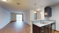 This single-floor residence offers a thoughtfully designed open-concept layout that seamlessly connects the kitchen and living areas. The kitchen is appointed with rich dark cabinetry, light granite countertops, a double stainless steel sink, and elegant pendant lighting that adds a contemporary touch. A half-wall peninsula serves as both a functional workspace and a natural divider between the kitchen and the open living space beyond. Throughout the unit, warm-toned hardwood flooring creates a cohesive and inviting atmosphere, complementing the clean white walls and neutral palette. The living area is bathed in natural light courtesy of a large sliding glass door fitted with vertical blinds, which leads to an outdoor area and enhances the sense of openness. Recessed and flush-mount ceiling fixtures provide ample ambient lighting throughout. Wood-trim accents around the doorways and baseboards add a subtle warmth to the overall aesthetic. The space presents a clean, modern ambiance with a practical and well-connected floor plan suited for comfortable everyday living.