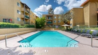 Located at 16830 Kingsbury Street in the Granada Hills neighborhood of Los Angeles, this outdoor amenity space serves as the central gathering hub of a multi-story residential community. The complex features warm golden-yellow stucco exteriors with distinctive Spanish-style red clay tile roofing accents, arched architectural details, and private balconies that overlook the courtyard below.

At the heart of the space is a generously sized rectangular swimming pool with clear, blue-green water, accessible via stainless steel handrails and steps. The surrounding pool deck is finished with light-toned tile pavers and is outfitted with multiple wicker-style lounge chairs, ideal for relaxing in the Southern California sun. A shaded seating area with an umbrella and bistro-style furniture provides a comfortable retreat alongside the deck.

The courtyard is framed by lush, mature landscaping including trees, shrubs, and bird-of-paradise plants, adding vibrant greenery throughout the grounds. Gated iron fencing encloses the pool area and courtyard pathways, providing both security and an open, airy feel. The overall ambiance is resort-like, combining Mediterranean architectural character with well-maintained outdoor living spaces designed for comfort and leisure.