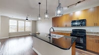 Located at 201 Town Center Ln in Keller, TX, this single-floor residence offers a thoughtfully designed open-concept layout with a seamless flow between the kitchen and living areas. The kitchen serves as the heart of the space, showcasing a large center island topped with dark granite countertops and a double-basin sink with a matte black faucet. Warm-toned wood cabinetry lines the walls, complemented by granite countertops, black appliances including a range, over-the-range microwave, refrigerator, and dishwasher. A trio of modern geometric pendant lights with an industrial flair hang above the island, adding a striking visual element. Track lighting provides additional illumination throughout the kitchen.

Rich hardwood flooring extends across the main living areas, creating a warm and cohesive aesthetic, while carpeting transitions into the bedroom spaces. The living area features a gently curved accent wall that adds an architectural touch, along with a ceiling fan. A well-lit hallway connects the private areas of the home, with natural light filtering in through large windows dressed with vertical blinds. The bedroom offers a bright, open feel thanks to a wide bank of windows and a ceiling fan overhead, making this a comfortable and functional residence.