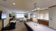 This single-floor amenity space is thoughtfully designed to offer a dynamic recreational environment. At its centerpiece sits a full-sized pool table, illuminated by a stylish industrial-style pendant light fixture that anchors the room with a modern yet inviting aesthetic. The patterned carpet adds visual interest underfoot and helps define the open layout.

A generous lounge seating area occupies one side of the room, featuring a sectional sofa and an upholstered ottoman, all arranged to encourage conversation and relaxation. Large windows with roller shades allow natural light to fill the space while offering privacy as needed. Potted indoor plants add a touch of greenery and warmth to the neutral color palette of soft whites, taupes, and warm wood tones.

Along one wall, a wet bar area with dark cabinetry and countertop provides a convenient station for refreshments, making this space well-suited for gatherings and events. Framed artwork lines the walls, contributing a polished, curated feel. Recessed ceiling lighting supplements the natural light throughout the space. An additional gaming station is visible near the entrance, further enhancing the recreational offerings of this versatile and well-equipped communal lounge.
