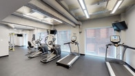 This fitness center offers a comprehensive workout environment designed to accommodate a wide range of exercise routines. The space features an industrial aesthetic, with exposed concrete beams and ductwork overhead, complemented by bright linear fluorescent lighting that ensures excellent visibility throughout.

The cardio zone is lined with large windows that allow natural light to filter in and offer views of the surrounding brick exterior architecture, creating an open and energizing atmosphere. This area is equipped with multiple treadmills, elliptical trainers, and stationary bikes, each outfitted with individual monitors for entertainment during workouts. Motivational phrases are displayed on the walls, adding to the encouraging environment.

The strength training section provides a well-rounded selection of equipment, including a full dumbbell rack with varying weights, a multi-function cable and weight machine, an adjustable workout bench, and medicine balls. A mirrored wall in this zone allows users to monitor their form during exercises.

The flooring throughout is durable dark rubber, ideal for high-impact activity and equipment use. Safety features such as a fire extinguisher are also present. The single-floor layout is efficiently organized, offering distinct zones for cardio and strength training while maintaining an open, spacious feel.