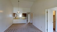 Located at 2100 Country Club Rd in Jacksonville, NC, this single-story home offers an open and airy layout designed for comfortable everyday living. The interior is defined by soaring vaulted ceilings that lend a sense of generous vertical space to the main living area, enhancing the overall openness of the floor plan. The living room is finished with wall-to-wall carpet in a neutral tone, providing a warm and inviting atmosphere throughout. A striking pendant light fixture hangs above the kitchen peninsula, serving as a functional and visual anchor between the living and kitchen areas. The kitchen features warm wood-toned cabinetry on both upper and lower sections, a built-in sink along the peninsula countertop, and a window above that allows natural light to filter into the space. The peninsula itself offers a convenient breakfast bar or prep area, seamlessly connecting the kitchen to the living space for an open, social feel. White walls throughout the home create a clean, bright backdrop that complements the natural wood cabinetry tones. A hallway visible from the main living area leads to additional rooms, suggesting a thoughtful and functional floor plan. Overall, the home presents a straightforward, well-proportioned single-level layout suited for a variety of lifestyles.