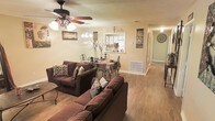 This single-floor home offers a comfortable and functional layout centered around an open-concept living and dining area. The living room is generously sized, accommodating two large sofas and providing an inviting atmosphere with warm, neutral-toned walls and durable hardwood-style flooring that runs throughout the main living spaces. A ceiling fan with integrated lighting fixture keeps the space comfortable year-round, while a chandelier above the dining area adds a touch of elegance. The dining area features a classic wrought-iron table set, seamlessly connected to the living space for easy entertaining. A charming pass-through window opens directly into the kitchen, enhancing the open feel and making it easy to interact between rooms. Decorative wall art and tasteful accents add personality throughout the space. A hallway visible from the main living area leads to additional rooms, suggesting a practical and well-organized floor plan. The overall ambiance is warm and homey, with cohesive decor and thoughtful use of space creating a relaxed, lived-in atmosphere that is both functional and inviting for everyday living.