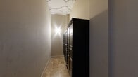 This single-floor hallway serves as a dedicated mail and package collection area for a residential building. The space is efficiently designed to maximize functionality within a compact footprint, with the majority of one wall lined by a large bank of numbered black U.S. Mail tenant mailboxes, offering individual locked compartments for residents. An additional freestanding black mailbox unit is positioned at the far end of the hallway, further expanding mail storage capacity.

The flooring consists of warm-toned stone tiles laid in a varied pattern, lending a sense of understated elegance to the utilitarian space. The ceiling is a notable architectural highlight, featuring decorative white molding with an ornate geometric and floral motif that adds a classic, refined character to the corridor. Smooth, neutral-toned walls keep the space feeling clean and uncluttered.

Lighting is provided by a wall-mounted sconce that casts a soft, upward and downward glow, creating a warm and welcoming ambiance throughout the narrow corridor. A wall-mounted HVAC unit ensures climate comfort. A resident notice is posted on the wall, reflecting the space's active use as a communal building amenity. Overall, the hallway balances practical design with thoughtful architectural details.