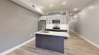 This thoughtfully designed one-bedroom, two-bathroom unit offers a contemporary living experience across a single, efficiently organized floor. The layout flows from a long, well-lit entry corridor finished with light wood-tone flooring and recessed lighting into a combined kitchen and living area that serves as the heart of the space.

The kitchen is a standout feature, equipped with stainless steel appliances including a refrigerator, electric range, and microwave. Two-tone cabinetry pairs warm wood-grain lower cabinets with light upper cabinets, complemented by white quartz countertops and a subway tile backsplash. A central island with an integrated sink adds both workspace and a casual dining option.

The living area opens directly onto a private balcony through floor-to-ceiling sliding glass doors, offering urban city views and abundant natural light. The bedroom is a compact, carpeted room with neutral gray walls and a transom-style window. A dedicated closet provides additional storage.

Both bathrooms maintain the unit's modern aesthetic, featuring large-format light tile flooring, dark wood-grain floating vanities, vessel sinks, illuminated mirrors, and frosted glass doors. Neutral gray walls and clean white trim unify the overall palette throughout the unit.