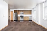 This single-floor space offers a clean, contemporary design with a thoughtful open-concept layout. The kitchen is a standout feature, outfitted with stainless steel appliances including a refrigerator, electric range, and built-in microwave. Warm wood-grain cabinetry lines the walls from floor to ceiling, complemented by granite-style countertops, a stainless steel sink, and a dishwasher. Rich hardwood-style flooring runs throughout the kitchen and main living areas, adding warmth and continuity to the space. Track lighting along the ceiling provides adjustable illumination throughout the main corridor and living zones. The bedrooms are carpeted in deep, dark tones and feature bright white walls, light wood baseboard trim, and windows fitted with horizontal blinds that invite natural light while maintaining privacy. Each bedroom offers generous proportions with views to the surrounding exterior. Storage is well addressed with built-in closets featuring light wood sliding doors. The bathroom is finished with a clean, minimal aesthetic — tile flooring, a soaking tub with shower, a wood-framed vanity with granite countertop, and chrome fixtures and hardware throughout. Overall, the space presents a cohesive modern design with functional finishes suited for comfortable everyday living.
