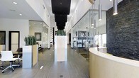 This single-floor commercial space showcases a sophisticated, modern design with a strong emphasis on transparency and open sightlines. The central corridor leads visitors past floor-to-ceiling glass-partitioned offices on both sides, allowing natural light to flow freely throughout the interior while maintaining a sense of visual connectivity between spaces.

The reception area anchors the space with a curved, light wood desk topped with a crisp white surface, creating a welcoming yet professional first impression. Overhead, a series of sleek pendant lights with metallic cylindrical shades hang at varying heights along the corridor, complementing a bold black ceiling panel that adds dramatic contrast to the otherwise bright, white palette.

Tall, sculptural planters housing snake plants are strategically placed throughout, introducing organic elements that soften the clean architectural lines. The flooring features linear gray tile that runs the length of the corridor, reinforcing a sense of depth and direction.

The glass-enclosed offices are furnished with modern workstations and task chairs, maintaining a professional and functional atmosphere. Dark mosaic tile accents on select wall sections add texture and visual interest, balancing the minimalist aesthetic with richness and depth throughout this polished commercial environment.