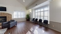 This single-floor space is designed as a versatile common or gathering area with a thoughtful, open layout that accommodates multiple functional zones. Vaulted ceilings lend an airy sense of height, while large windows fitted with horizontal blinds allow generous natural light to filter across the rich, wood-look flooring that runs throughout the entire space.

The main lounge area is anchored by a striking brick fireplace with an arched surround, complemented by a wall-mounted television and leather seating. Adjacent to this is a dedicated waiting or seating zone featuring a row of sleek black chairs positioned along the windows, ideal for gatherings or organized use.

A round dining table with black upholstered chairs occupies a defined corner, illuminated by a modern pendant chandelier. Decorative wall art and a mirror arrangement add visual interest to the neutral, wainscoted walls. A cozy secondary seating nook with patterned accent chairs sits near a pass-through opening to the galley-style kitchen, which is equipped with stainless steel appliances, a gas range, built-in microwave, and white cabinetry with light countertops. The overall ambiance is clean, functional, and welcoming.