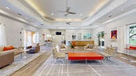 Located at 2450 Lake Debra Drive in Orlando, Florida, this single-floor community clubhouse lounge offers a generously sized, open-plan living area designed for comfort and social interaction. The space is defined by its striking tray ceilings with recessed lighting and ceiling fans, which lend a sense of architectural depth and airiness throughout. Wood-look tile flooring runs seamlessly across the majority of the space, transitioning to plush area rugs in designated seating zones that help define distinct gathering areas within the open layout. Large sliding glass doors and floor-to-ceiling windows flood the interior with natural light while offering direct views and access to the surrounding outdoor courtyard. The main living area is furnished with multiple seating arrangements, including neutral-toned sofas and armchairs accented with orange throw pillows, a bold orange bench as a central focal point, and round accent tables that encourage conversation. A built-in credenza with wood cabinetry provides functional counter space along one wall, accompanied by vibrant framed artwork that adds color and personality throughout. A dedicated lounge alcove features a wall-mounted television flanked by colorful art panels, creating an intimate media nook. The overall ambiance is bright, modern, and welcoming, combining contemporary design elements with warm, inviting furnishings.
