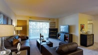 This single-floor residence centers around a comfortable living room designed for both relaxation and functionality. The space is anchored by a dark-tiled fireplace, which serves as a striking focal point on one wall, adding warmth and character to the room. Adjacent to the fireplace, a media console houses a flat-screen television, providing a practical entertainment setup.

Large sliding glass doors dominate one wall, flooding the living area with natural light and offering views of the surrounding greenery outside. Vertical blinds offer privacy and light control as needed. The warm yellow-toned walls complement the neutral carpeted flooring, creating an inviting atmosphere throughout.

The living room is furnished with dark leather sofas arranged around a rectangular coffee table, providing ample seating for guests. Decorative accents, including a sculptural horse figurine and a textured wall piece, add personality to the space. Built-in ventilation is visible in the ceiling, ensuring comfortable climate control year-round. A door on the left side of the room leads to additional areas of the residence, suggesting a practical and connected layout.