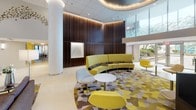 Located at 8560 Second Ave in Silver Spring, Maryland, this single-floor commercial lobby makes a striking first impression through a thoughtful blend of contemporary design elements and vibrant accents. The space is anchored by a bold geometric-patterned carpet in yellow, gray, and brown tones that energizes the floor and visually defines multiple seating clusters throughout the area.

A standout crystal chandelier suspended from a coffered ceiling serves as a luminous centerpiece, complemented by recessed cove lighting that casts a warm, ambient glow across the space. The ceiling itself features tiered architectural detailing that adds depth and a sense of grandeur to the open layout.

Rich, dark wood-paneled walls provide a sophisticated contrast to the bright furnishings and light-toned flooring. A curved, chartreuse sectional sofa anchors one seating zone, while modern pod-style chairs in yellow, gray, and white are arranged around sleek round tables to create inviting conversation areas. Tall glazed panels allow natural light to filter in and offer views of the surrounding outdoor greenery, maintaining a connection to the exterior environment.

A marble reception desk is positioned prominently near the entrance, providing a polished focal point for visitors. Decorative wall art and subtle sculptural accents further enrich the interior, contributing to a professional yet welcoming ambiance throughout this well-appointed lobby.