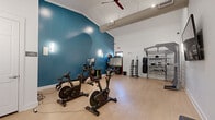 This dedicated fitness studio offers a thoughtfully organized workout space spread across a single open floor. The room is defined by a striking teal accent wall that contrasts with the surrounding white walls, creating an energetic and motivating atmosphere. Vaulted ceilings with exposed ductwork and a ceiling fan add a sense of vertical openness, while wall-mounted sconces provide warm, ambient lighting throughout.

The light wood-look flooring runs throughout the space, offering a clean and durable surface suited for a variety of exercise activities. Cardio equipment includes multiple stationary cycling bikes with integrated display screens and a rowing machine, all positioned to allow comfortable use and spacing. A multi-functional training rack with integrated weight storage, pull-up bars, and a heavy bag attachment anchors one side of the room for strength and functional training. A dumbbell rack stocked with a full range of weights provides additional resistance training options.

A wall-mounted flat-screen television serves as a focal point on the white end wall, ideal for guided workouts or entertainment. A dedicated yoga storage room is accessible via a separate door, keeping the main floor clear and organized. The space is efficiently ventilated with visible HVAC vents and benefits from natural light through a window near the exit door, completing a clean, functional, and well-appointed fitness environment.