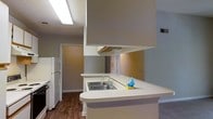 This single-floor residential space is thoughtfully laid out to maximize functionality and flow. The kitchen serves as the heart of the unit, designed with a peninsula-style counter that extends outward to create a natural divide between the kitchen and the adjoining living area. The countertops are finished in a clean white laminate surface, offering ample workspace, while a stainless steel sink is integrated into the peninsula for added convenience. White cabinetry lines the kitchen, providing generous storage, and is complemented by an under-cabinet range hood positioned above the cooking area. A full-size white refrigerator anchors one side of the kitchen. The flooring throughout the main areas features warm-toned hardwood-style planks, adding a grounded, cohesive aesthetic to the space. The adjoining rooms are finished with neutral-toned walls and carpeting, creating a calm and functional living environment. A built-in closet with bi-fold doors is visible in the background, adding practical storage to the living or bedroom areas. Overhead fluorescent lighting ensures the space is well-lit throughout. The overall design is clean, practical, and well-suited for everyday living.