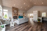 This single-floor commercial space blends professional functionality with warm, inviting design. The centerpiece is a spacious great room boasting soaring vaulted ceilings clad in natural wood planking, complemented by rich hardwood-style flooring throughout. A floor-to-ceiling stacked stone fireplace anchors the room, topped with a large decorative clock and a rustic wood mantel, creating a standout focal point. Generous windows along the front façade flood the space with abundant natural light while offering open views of the surrounding landscape. The reception and lounge area near the entrance features high ceilings with large multi-pane windows, providing a bright and welcoming atmosphere for visitors. A kitchenette area offers full-height gray shaker cabinetry, granite countertops, a sink, and countertop appliances, ensuring convenience for daily use. A dedicated conference room accommodates a full-sized conference table and ergonomic seating, with a window providing natural light and a serene exterior view. Private office areas are accessible via glass-panel doors, featuring ample workspace, large windows, and open, airy layouts. The overall ambiance is cohesive and thoughtfully appointed, balancing comfort and professionalism throughout.