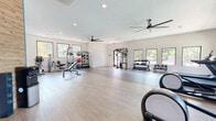This fitness center is designed to support a comprehensive workout experience within a bright, open, and well-organized single-floor layout. The space features light wood-style flooring with subtle accent stripes that define different workout zones, contributing to a clean and modern aesthetic throughout.

The main workout area is generously proportioned, offering ample floor space for free movement and functional training. Two large ceiling fans ensure consistent air circulation across the room, complementing the recessed lighting overhead. Large windows along multiple walls invite abundant natural light and provide views of the surrounding greenery, creating an energizing atmosphere.

Equipment is thoughtfully arranged throughout the space. A dedicated free weights section includes a full rack of dumbbells in graduated sizes. A multi-function cable and resistance training station serves as a central strength-training anchor. A well-stocked ball rack holds a variety of medicine balls and stability balls for dynamic and functional workouts. Additional strength machines and a weight bench are positioned along one side, accompanied by a floor-length mirror wall that enhances the sense of openness.

The entry area features a distinctive exposed brick accent wall alongside double glass doors, a water dispenser, and foam rollers for warm-up and recovery. The facility also includes cardio equipment such as treadmills and an elliptical machine, rounding out a complete fitness offering.