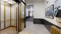 Located at 1245 Rue St-Marc in Montréal's H3H neighbourhood, this hallway offers a refined glimpse into classic Montreal residential architecture. The corridor is defined by striking dark marble wainscoting that lines both walls, creating a bold contrast against the crisp white upper walls and an ornate embossed ceiling. A central runner of grey carpet guides visitors along the length of the space, flanked by light tile flooring on either side.

Rich, dark-stained hardwood door frames and unit doors punctuate the hallway at regular intervals, lending a sense of traditional craftsmanship throughout. Elegant globe pendant light fixtures hang from the ceiling, providing warm, even illumination that highlights the carefully preserved architectural details.

At one end, a beautifully crafted entry door featuring a vibrant stained glass transom window serves as a focal point, showcasing a floral motif in rich reds and greens — a hallmark of early 20th-century decorative artistry. A vintage elevator with polished brass finishes adds further character near one end of the corridor.

The combination of heritage materials, classic detailing, and thoughtful lighting creates an atmosphere of timeless elegance, making this hallway a distinguished and welcoming introduction to the building.
