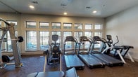 This fitness center is designed to support a comprehensive workout experience within a spacious, single-level layout. The cardio zone is positioned along a wall of large, multi-pane windows that fill the space with natural light and provide views of an outdoor patio and surrounding grounds. Multiple treadmills, an elliptical, a stair climber, and a Cybex cross-trainer are arranged to take full advantage of this bright setting, creating an energizing environment for aerobic exercise.

The strength training area occupies the interior of the space and features a multi-function cable machine, a free weight rack with dumbbells, an adjustable bench, and additional resistance equipment. Structural columns are integrated into the layout and are flanked by floor-to-ceiling mirrors that visually expand the room and allow users to monitor their form throughout their workout. A wall-mounted flat-screen television is positioned above the strength equipment, adding an entertainment element to the space.

Flooring transitions between carpet in the cardio zone and wood-style hard flooring in the strength area. Recessed lighting throughout ensures consistent illumination. A textured accent wall and open shelving add subtle design character. The overall ambiance is clean, modern, and functional.