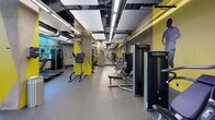 Located at 1630 Chicago Ave in Evanston, Illinois, this fitness center offers a well-appointed, single-floor workout facility designed with a bold industrial aesthetic. Exposed ductwork, open ceilings, and concrete structural elements are complemented by striking yellow accent walls and columns, creating an energetic and motivating atmosphere throughout the space.

The strength training area is equipped with a comprehensive selection of cable machines, selectorized weight machines, and free weights, including a full dumbbell rack with an adjustable bench. Rubber flooring provides safety and durability in this zone. A dedicated cardio section features treadmills and additional cardio machines, positioned near large windows that frame views of the surrounding Evanston cityscape, filling the space with natural light.

A partitioned cycling studio houses multiple stationary bikes, with large street-facing windows offering an open, airy feel during rides. Adjacent to this is a flexible studio area finished with wood-look flooring, perforated metal room dividers, and wall-mounted instructional displays, making it ideal for stretching, yoga, or guided workouts. A water fountain station is conveniently located within the main floor. The layout flows openly across all zones, supporting a full range of fitness activities in a cohesive, modern environment.