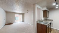This single-story residence offers a practical and comfortable floor plan across two bedrooms, two bathrooms, and a combined kitchen and living area. The open living room features soft wall-to-wall carpeting, neutral beige tones, a textured ceiling, and sliding glass doors that lead to a private balcony, allowing natural light to fill the space and providing pleasant outdoor views. A front entryway with hardwood-style flooring transitions seamlessly into the carpeted living area.

The kitchen is positioned adjacent to the living room and features warm wood-toned cabinetry, granite-style countertops, a double-basin sink, track lighting, and hardwood-style flooring. A black refrigerator anchors the far end, and a dedicated pantry closet with built-in shelving offers generous storage.

The primary bedroom is a comfortable retreat, complete with plush carpeting, a ceiling fan with light fixture, and a direct en-suite bathroom access. The second bedroom also features carpet flooring, a closet, and a window for natural light. Both bathrooms are fitted with full tub-shower combinations accented by clean white tile surrounds. A hallway connects the bedrooms and bathrooms, with an additional closet providing extra storage throughout the home.