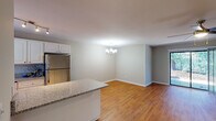 This single-floor residence in South Carolina offers a cohesive and functional layout with warm hardwood-style flooring that flows consistently throughout every room. The open-concept design seamlessly connects the kitchen, dining, and living areas, creating a spacious and inviting atmosphere. The kitchen is well-equipped with white cabinetry, granite countertops, stainless steel appliances including a full-size refrigerator, a built-in range, and track lighting overhead. A breakfast bar provides an additional casual dining option. The dining area features an elegant chandelier, adding a refined touch to the space. The living room is bright and airy, highlighted by a ceiling fan and sliding glass doors that open to the outdoors, providing pleasant views of surrounding greenery. Multiple bedrooms offer comfortable proportions, each fitted with ceiling fans and windows that invite natural light. The primary bedroom connects to a private bathroom featuring white cabinetry, granite countertops, a large mirror, and stylish vanity lighting. A second bathroom provides a granite-topped vanity and a shower-tub combination. Neutral gray tones on the walls and crisp white trim throughout give the home a clean, move-in-ready appeal.