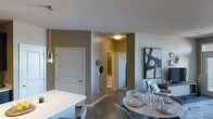 This single-floor residence showcases a thoughtfully designed open-concept layout that connects the dining and living areas with ease. The dining space is anchored by a round table with woven placemats and contemporary place settings, surrounded by modern upholstered chairs that add a touch of refinement. Sleek pendant lighting with metallic finishes hangs nearby, complementing the clean white surfaces of the adjacent kitchen counter. The polished concrete-style flooring runs throughout, providing a cohesive and low-maintenance foundation that enhances the modern aesthetic. Walls are finished in warm taupe and neutral grey tones, lending a calm, sophisticated ambiance to the space. A hallway visible in the background leads to additional rooms, including what appears to be a bathroom, offering a sense of depth and privacy within the layout. White panelled doors provide clean architectural contrast against the warm wall tones. Abstract wall art adds a curated, gallery-like quality to the dining area. Overall, the space balances contemporary design elements with functional living in a well-organized single-level floor plan.