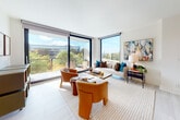 This thoughtfully designed single-floor residence combines modern architecture with warm, curated interiors. The open-plan living and dining area serves as the heart of the home, where expansive floor-to-ceiling windows and sliding glass doors flood the space with natural light and frame sweeping outdoor views of the surrounding treetops and skyline. A private balcony extends the living space further, accessible directly from the living room. The living area features light tile flooring, contemporary furnishings, and bold artwork, creating an inviting yet sophisticated ambiance. Adjacent to it, the dining area comfortably accommodates a large wood table and is accented by a brass flush-mount fixture and wall sconce. The galley-style kitchen is well-equipped with white cabinetry, gold hardware, stainless steel appliances, a gas range, and a light stone countertop with an undermount sink. The bedrooms offer serene retreats, each with large windows capturing natural light and views. The primary bedroom features ample space and direct access to additional rooms. Both bathrooms are finished with marble-tiled walk-in showers, white vanities with brass hardware, and light wood-look tile flooring, delivering a clean, polished aesthetic throughout.