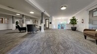 Located at 1701 Cedar Hill Cross Rd in Saanich, BC, this building's main-floor lobby and hallway offer a welcoming and refined common area that blends classic architectural character with modern interior finishing. The entry foyer is anchored by a striking two-tiered chandelier with exposed Edison-style bulbs, suspended beneath a decorative crown-moulded ceiling. Wainscoting panels line the walls, complementing the grey patterned carpet underfoot. Large windows and a cross-panelled front door flood the entry with natural light and offer views of lush greenery outside. A built-in mailbox bank and a sleek bench provide practical amenities at the entrance. The hallway opens into a generously proportioned common lounge area, where structural columns with classical detailing define the space. Textured ceiling tiles add architectural interest overhead, while flush-mount lighting fixtures maintain a clean, well-lit environment. The lounge is furnished with plush grey accent chairs and side tables, creating comfortable seating vignettes throughout. Potted fiddle-leaf fig trees introduce organic warmth. A staircase connects to additional areas of the building, and interior doors lead to further amenities. The overall ambiance is polished, calm, and inviting.