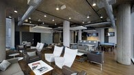 This single-floor space showcases a compelling blend of industrial and contemporary design. The exposed concrete ceiling, complete with visible ductwork and conduit, creates a raw, urban aesthetic that is complemented by recessed and track lighting, providing ample illumination throughout the open-plan layout. Structural columns punctuate the floor plan, adding to the industrial character while defining distinct seating zones.

The lounge area is furnished with a curated mix of high-back white chairs, dark brown leather armchairs, and a neutral-toned sofa arrangement, offering versatile seating configurations suited for both individual relaxation and group conversation. Low coffee tables and side tables are distributed throughout, adding functionality to the space.

Toward the rear, a bar or service counter area is visible, accompanied by a wall-mounted television, suggesting a social or entertainment-oriented function. Writable glass panels and wall-mounted artwork contribute to a creative, collaborative atmosphere. The warm-toned hardwood flooring anchors the space, providing a welcoming contrast to the cool, industrial overhead elements, resulting in an overall ambiance that feels both modern and inviting.