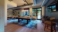 This thoughtfully designed single-floor communal space blends industrial aesthetics with modern comfort across several distinct functional areas. The kitchen is outfitted with rich, dark wood cabinetry extending floor to ceiling, providing generous storage, and is equipped with stainless steel appliances including a gas range, microwave, and dishwasher, complemented by dark stone countertops and light hardwood flooring. Adjacent living and lounge areas feature a sophisticated palette of deep navy walls, exposed concrete columns, and warm leather and upholstered furnishings, creating an inviting atmosphere for relaxation and socializing. A dedicated media room offers a large wall-mounted flat-screen television, a sound bar, and comfortable lounge seating arranged around a concrete coffee table, with roller shades providing light control. The standout billiards room showcases exposed ductwork and industrial ceiling details, a full-sized pool table centered on a patterned area rug, and large windows and a glass door opening directly to an outdoor patio with views of lush greenery. An ADA-compliant restroom features grab bars, a wall-mounted sink, and clean tile flooring, ensuring full accessibility. Together, these spaces offer a cohesive and versatile environment suited for both leisure and community engagement.